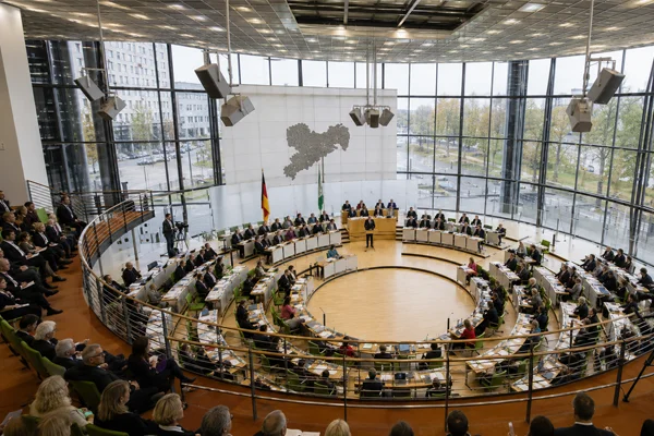 Plenarsaal des Sächsischen Landtags in Dresden während einer Sitzung: Abgeordnete sitzen im halbkreisförmigen Parlament unter hoher Glasdecke, Kameras begleiten die Debatte. Das Bild illustriert die politische Kontroverse um die sogenannte „Brandmauer“ nach einer Abstimmung, bei der ein Antrag der Grünen mit Stimmen von AfD und BSW eine Mehrheit erhielt.